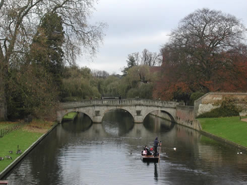 Small boat being steered down smooth, narrow waterway near a stone bridge on an overcast day